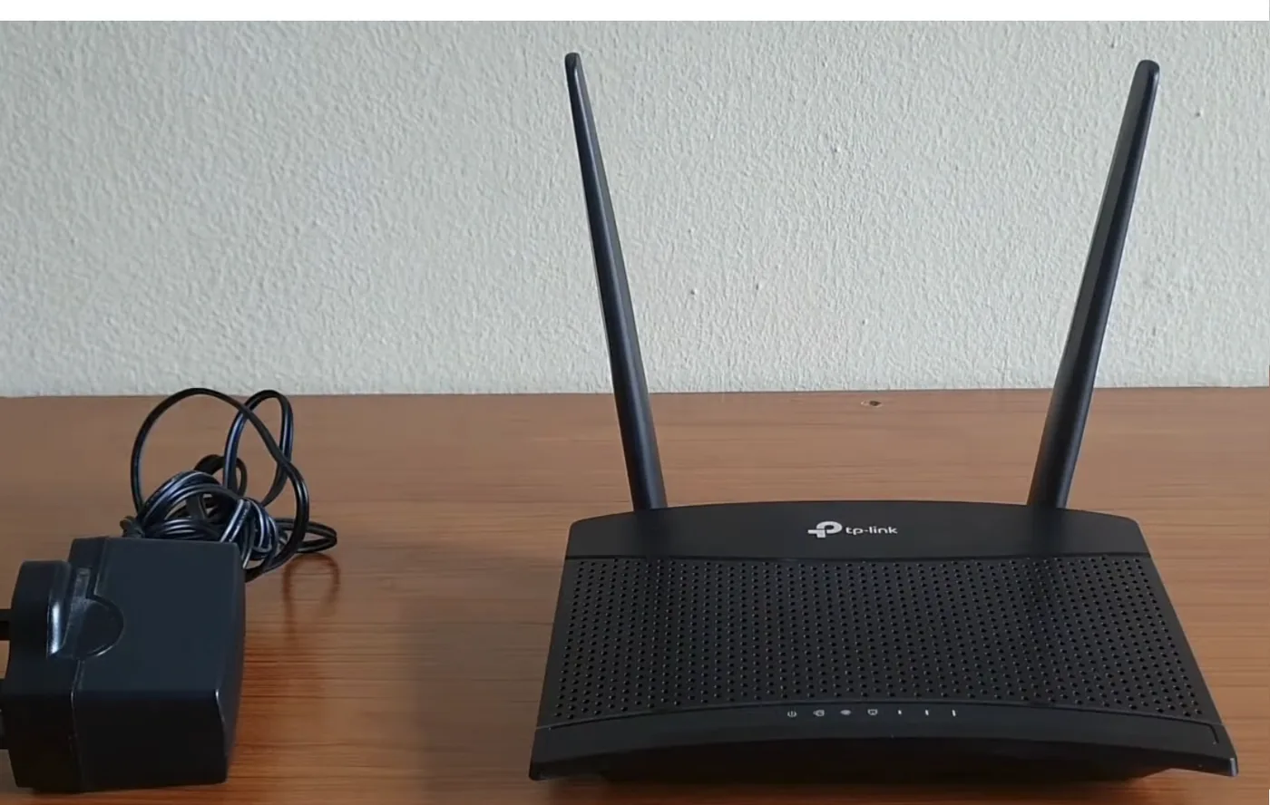 A black four gigabyte router with two upright external antennas sitting on a wooden desk against a pale wall, with its coiled power supply beside it. A row of status lights runs along the front edge of the case.