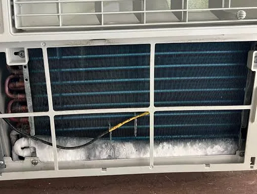 The front grille removed from a window air conditioner inside a cold room, showing the evaporator coil. A band of white ice has built up along the bottom third of the coil and across the pan below it. A sensor lead is taped across the coil face.
