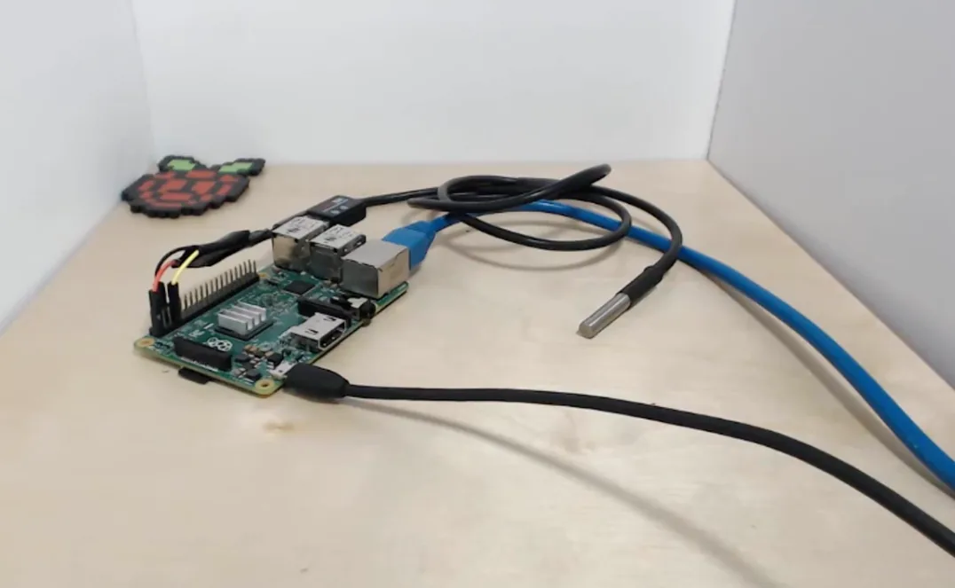 A bare Raspberry Pi board on a plywood bench with no case. Three jumper wires run from its pin header to a stainless temperature probe on a black lead, and an ethernet cable and a power lead are plugged in. A Raspberry Pi logo coaster sits behind it.