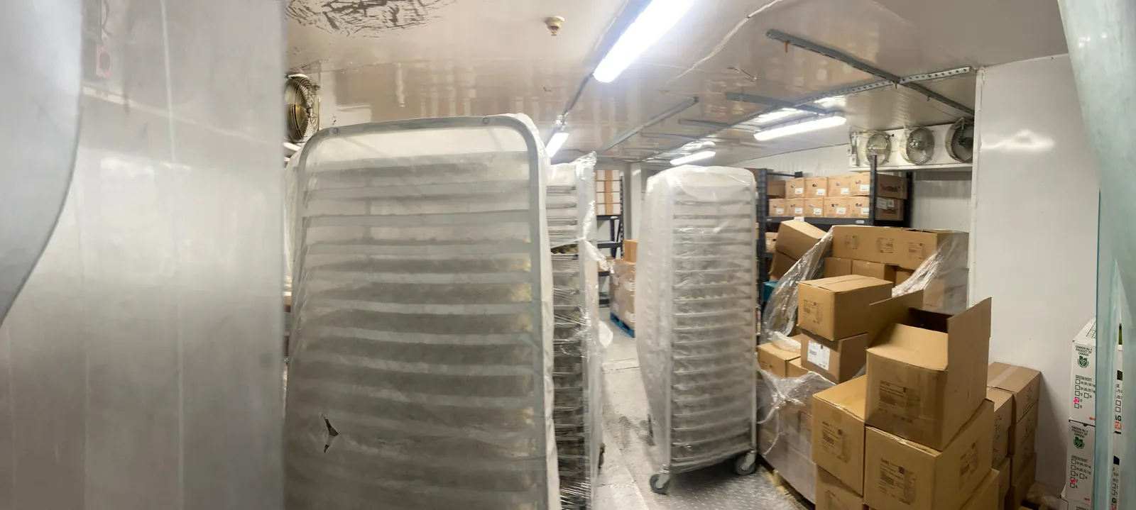The inside of a loaded walk-in cooler. The walls and ceiling are bare metal panels. Two sheet pan racks wrapped in plastic stand in the aisle, pallets and stacked cartons fill the far end, and a three fan evaporator coil is mounted high on the right hand wall.