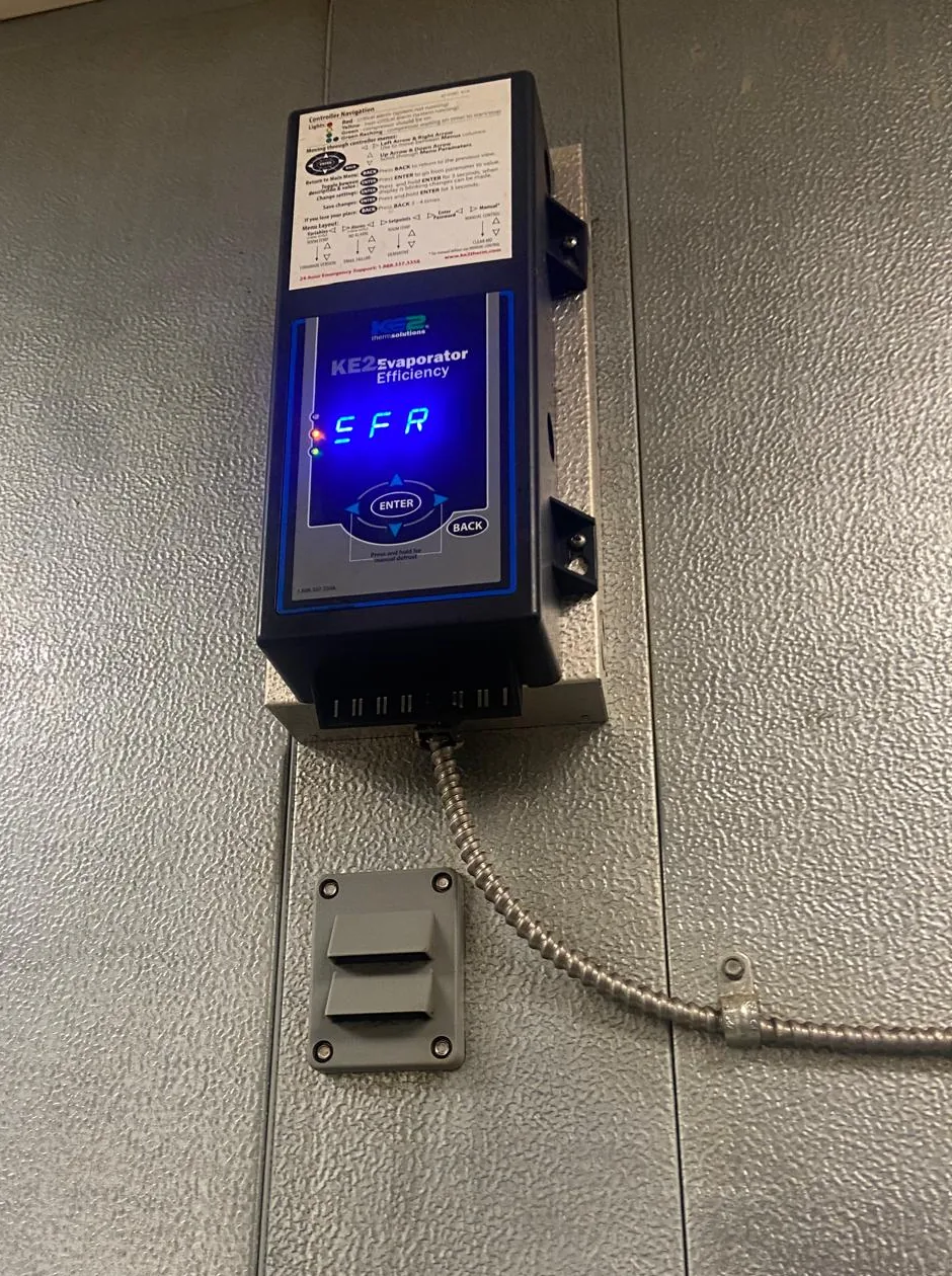 A refrigeration controller mounted on the insulated panel wall outside a walk-in. Its blue display is lit and showing a three character code, with red and green status lights beside it and a navigation reference card stuck to the case above. Flexible metal conduit runs from the base of the unit down and across the wall.