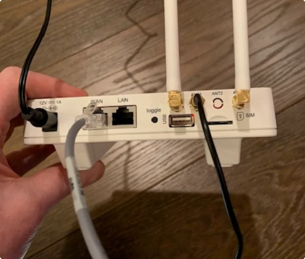 The back of a monitoring hub held in a hand. A grey Ethernet cable is plugged into the WAN port. The LAN port next to it is empty. A SIM slot sits on the right, beside two cellular antennas.