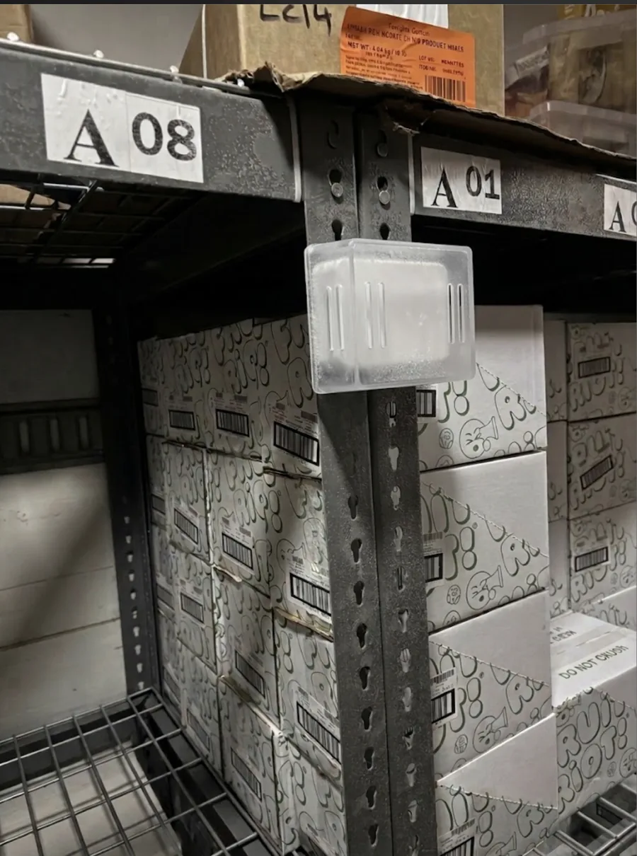 A wireless temperature sensor clipped to the upright of a pallet rack inside a loaded walk-in freezer, at about the height of the stock on the shelf. The case is covered in frost. Cartons of product are stacked on the shelf behind it and the rack locations A08 and A01 are labelled above.
