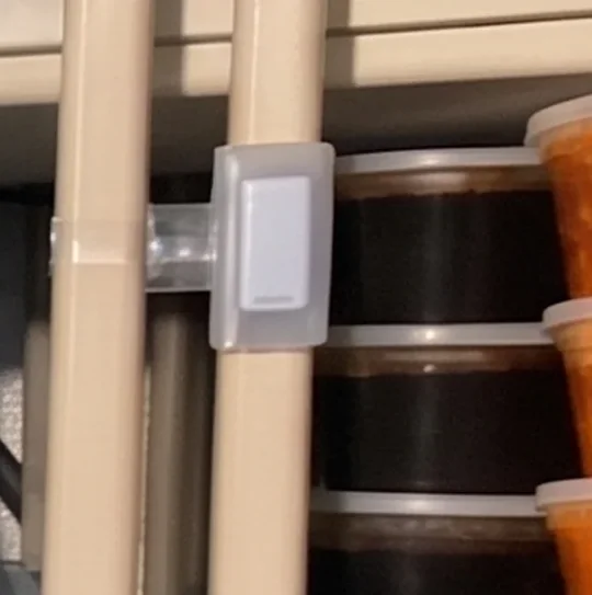 A close view of a TemperatureWise sensor in its bracket on a shelving upright inside a walk-in, with stacked food containers behind it.