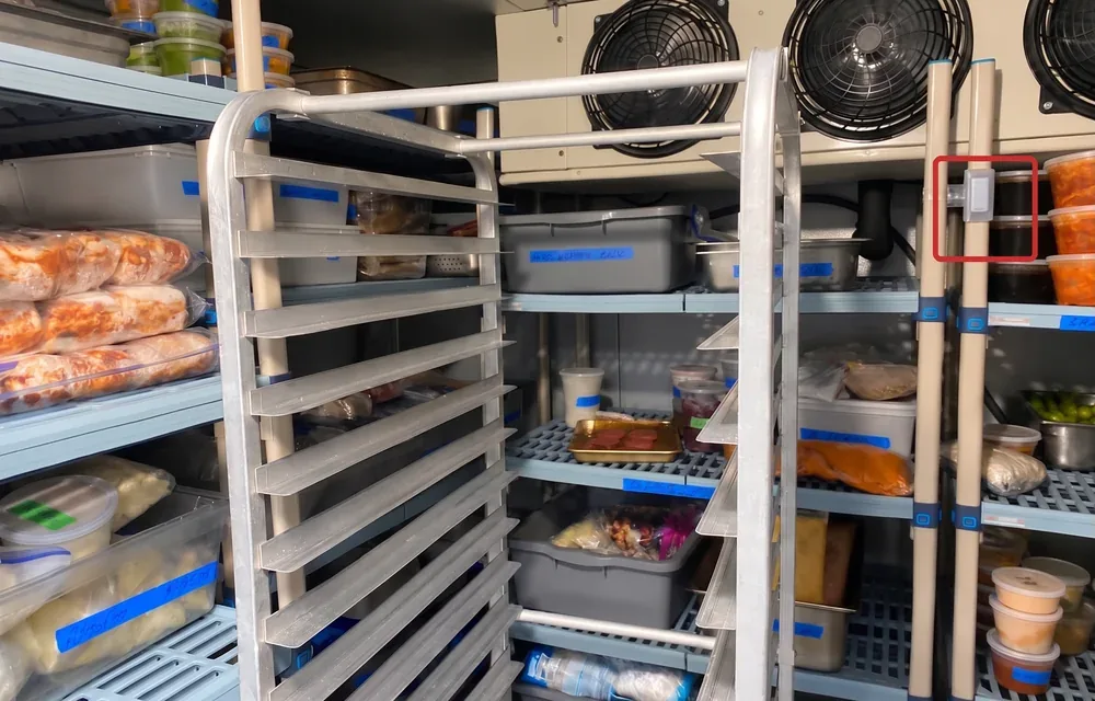 Inside a loaded walk-in cooler: wire shelving stacked with labelled food containers, a sheet pan rack, the evaporator fans above, and a small white TemperatureWise sensor clipped to a shelving upright, marked with a red outline.